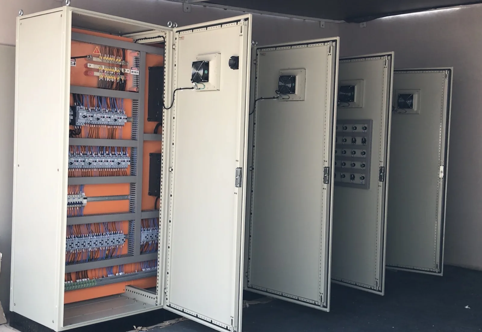 Fileira de painéis elétricos com porta aberta mostrando placa de componentes laranja com disjuntores e cabeamento